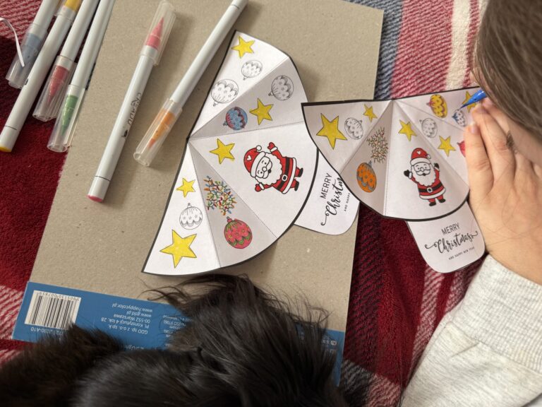 Dziecko koloruje i składa papierową świąteczną zabawkę z Mikołajem i bombkami. Na stole leżą flamastry, karton oraz gotowa i częściowo złożona papierowa forma; całość utrzymana w ciepłej, domowej atmosferze, bez użycia ekranów.